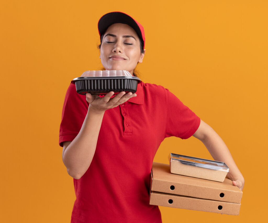 pleased young delivery girl wearing uniform and cap holding pizza boxes and sniffing food containers in her hand isolated on orange background